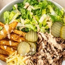 A colorful burger bowl with fresh vegetables, seasoned meat, and toppings served in a bowl.